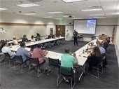 approximately 20 people sitting around long white tables set in a U-shape listening to DCTC Instructor Bob Trewentha with AI Instructor, Matt Boudinot in the background leaning on a brown podium. 